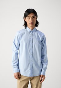 Chemise boutonnée bleu clair en tissu lisse, dotée d'un col classique, de manches longues et de détails de couture subtils.