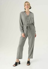 Grijze gemarmerde jumpsuit met kraag, knoopsluiting aan de voorkant, verstelbare tailleband, lange mouwen en omgeslagen enkels, gecombineerd met zwarte hakken.