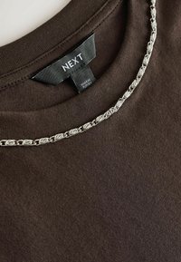 Smeđa pamučna majica s okruglim izrezom, s naglaskom na srebrni lanac duž ovratnika i crnom oznakom "NEXT" ušivenom iznutra.