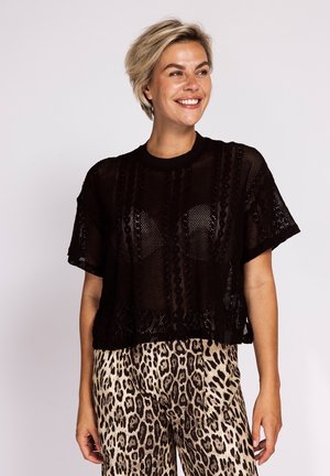 Smiling woman with short blonde hair wearing a black see-through knitted top and leopard print pants, standing against a plain background.