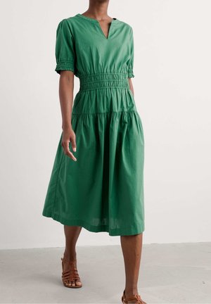 Woman wearing a green, knee-length dress with short puff sleeves and brown strappy sandals standing on a gray floor.