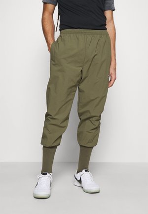 Personne portant un pantalon court vert olive avec des poignets élastiques, des chaussettes côtelées olive, des baskets blanches et un haut à manches courtes noir, debout contre un mur blanc.