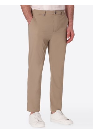 Pantaloni beige slim fit con chiusura con bottone e zip, indossati con sneakers bianche e camicia bianca infilata, mani nelle tasche.