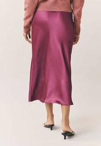 Magenta satenska midi krila z gladko teksturo, ki ima rahlo širjenje ob robu, v kombinaciji s črnimi sandali na peto.