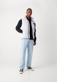 Gilet matelassé blanc avec un col montant, associé à une chemise noire à manches longues, un jean bleu clair et des baskets noires. Fond blanc.
