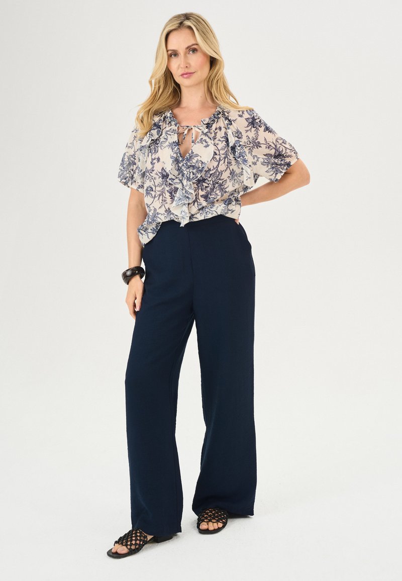 Blumige Bluse mit gerafftem Ausschnitt, kurzen Ärmeln und Schlüssellochdetail, kombiniert mit hochtaillierten, marineblauen Weitbein-Hosen und strukturierten schwarzen Sandalen.