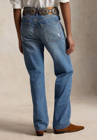 Osoba nosící modré džíny s rovným střihem, zadními kapsami, dekorativním hnědým pásem a hnědými botami s špičatou špičkou, stojící na neutrálním pozadí.