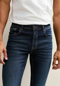 Man som bär mörkblå jeanstight och en vit t-shirt, står mot en ljus vägg.