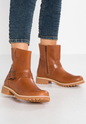 Nilkkaa peittävät vaaleanruskeat nahkasaappaat, joissa on sivuvetoketju ja solkikiristys, pidettynä tummansinisillä käännetyillä farkuilla valkoisella lattialla.