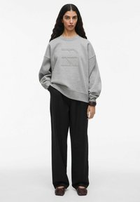 Felpa grigia con logo in rilievo, spalle scese e maniche ampie, abbinata a pantaloni neri a gamba larga e scarpe slip-on marroni.