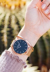 Reloj de esfera negra con banda de malla en oro rosa, con un diseño minimalista y delgadas marcas horarias. Llevado en una muñeca cubierta con un suéter claro.