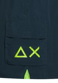 Il tessuto blu navy presenta una grande tasca frontale con simboli grafici in neon verde: un triangolo, un cerchio e una croce. Include un accento giallo neon alla base.