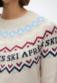 Beige kootud sviiter ümmarguse kaelusega, millel on sinised, punased ja mustad zikk-zakk mustrid ning südamekujulised aktsendid, samuti tekst "SKI APRÈS."