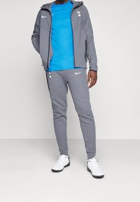 Giacca grigia con zip e cappuccio, t-shirt blu e pantaloni jogger grigi, tutti con il logo Nike; scarpe sportive bianche, texture del tessuto liscia.