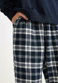 Die Hand einer Person ist in der Tasche, die navyblaue und weiße karierte Hose und einen navyblauen Pullover trägt, zu sehen von der Mitte des Oberkörpers bis zum Oberschenkel.