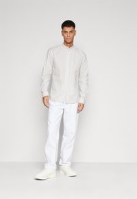 Button-up shirt with light beige and white vertical stripes, mandarin collar, long sleeves, paired with white pants and white sneakers.