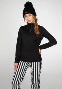 Sudadera de forro polar negra con cuello alto y cremallera, combinada con pantalones a rayas negros y blancos. Modelo luciendo un gorro de lana negro con pom-pom.