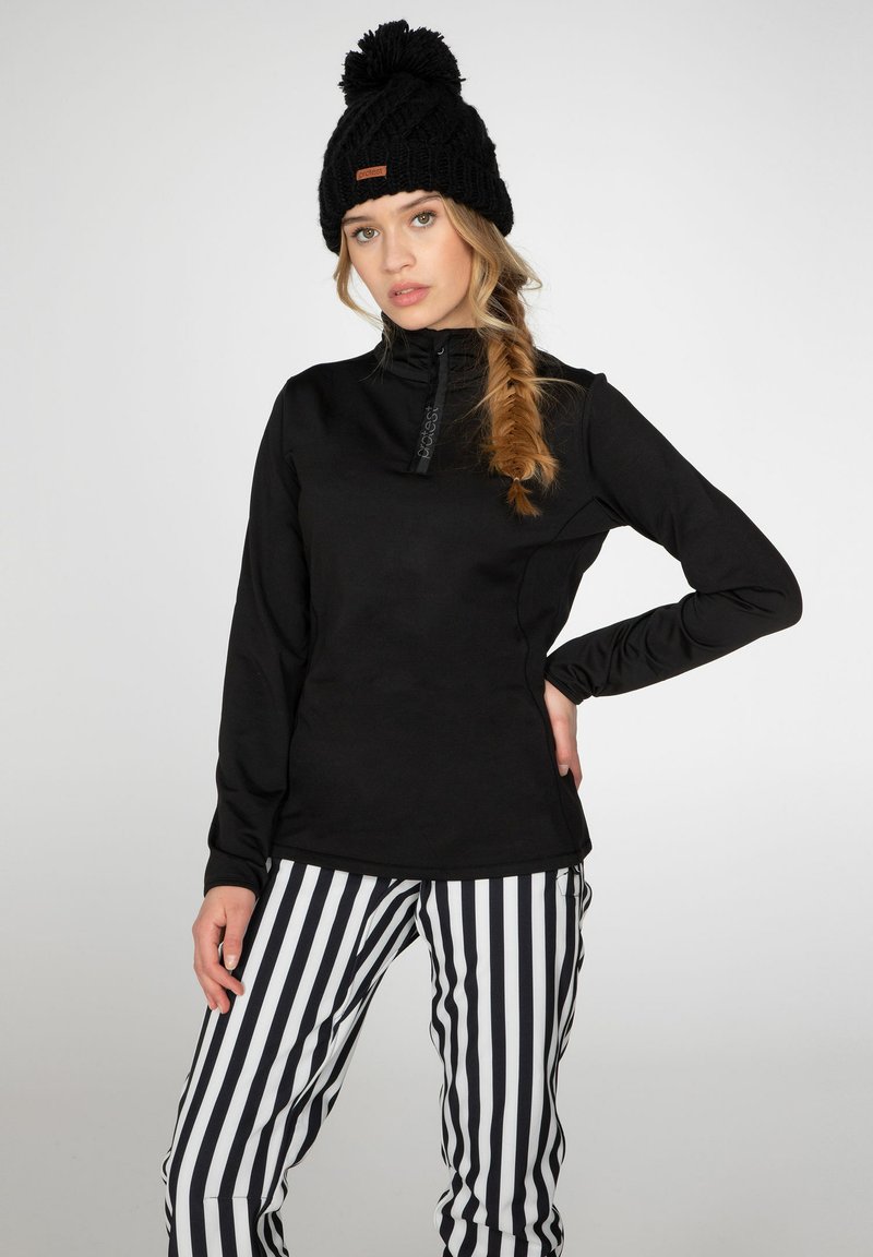 Sudadera de forro polar negra con cuello alto y cremallera, combinada con pantalones a rayas negros y blancos. Modelo luciendo un gorro de lana negro con pom-pom.