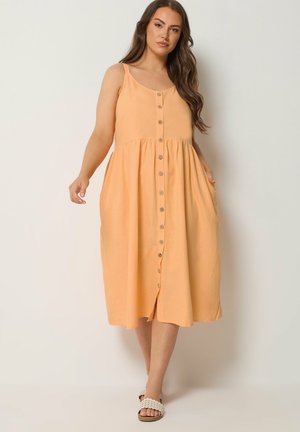 Kvinde iført en ærmeløs, lyserød midi-kjole med knapper foran og sidelommer, stående mod en ensfarvet lys baggrund.