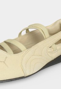 Chaussure plate beige avec semelle noire, brides élastiques sur le dessus et logo Puma cousu sur le côté, conçue pour un usage décontracté.