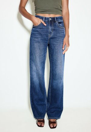 Kvinde iført blå jeans med høj talje og vide ben, grøn cropped top og brune åbne hæle, stående med den ene hånd i lommen.