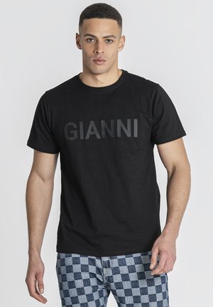 Muž v čiernom tričku s nápisom "GIANNI" a modro-sivo kockovaných nohaviciach, stojaci na jednoduchom pozadí.