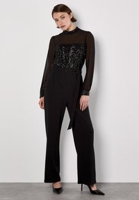 Schwarzer Jumpsuit mit einem pailettenbesetzten Oberteil, transparenten Ärmeln und einem taillierten Bund. Der Stoff hat eine glatte Textur und ein weites Bein-Design. Schwarze Absätze.