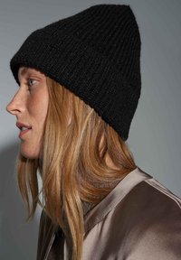 Zwarte gebreide beanie met een geribbelde textuur, taperend naar de bovenkant, gedragen met een lichtbruin satijnen jack. Heeft een casual pasvorm.