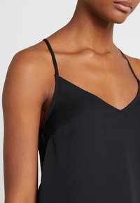Top camicia nera con spalline sottili, design a V, tessuto morbido e leggermente lucido. Le cuciture sui lati esaltano la forma e la vestibilità.