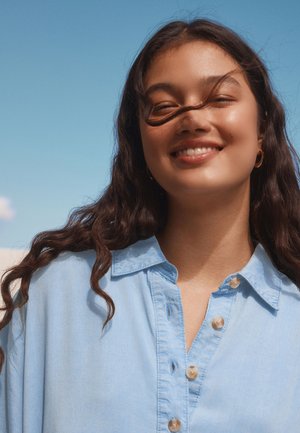 Jauna sieviete ar gariem viļņainiem matiem, tērpusies gaiši zilā pogājamas kreklā, smaidot ārā pret skaidru zilu debesi.