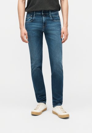 Personne portant un jean bleu slim et des baskets blanches à lacets, debout devant un fond clair uni.