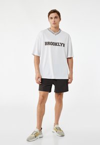 Jersey blanco de manga corta con un cuello de rayas negras y el texto "BROOKLYN". Se lleva con pantalones cortos negros y zapatillas de color claro.