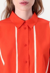 Camicia rossa con bottoni, caratterizzata da un colletto classico, strisce bianche verticali e una trama di tessuto liscia. Cinque bottoni visibili sul davanti.