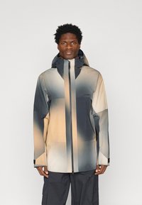 Man wearing a hooded, zip-up jacket with beige, gray, and black gradient pattern and black pants, standing against plain background.