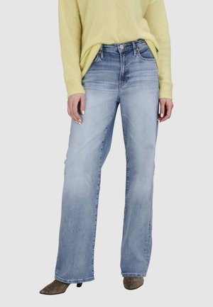 Femme portant un jean à taille haute et jambes larges de couleur bleu clair, un pull jaune et des chaussures marron à bout pointu, debout avec les mains détendues le long du corps.