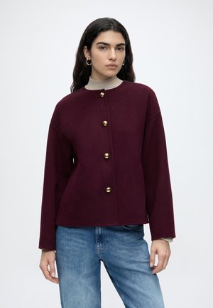 Femme aux longs cheveux foncés portant des boucles d’oreilles créoles dorées, une veste bordeaux boutonnée, un col roulé beige et un jean bleu sur fond blanc.