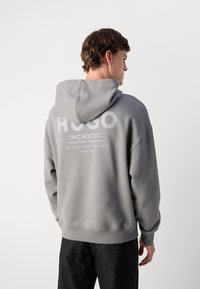 Grå hettegenser med snøring, med en dristig hvit "HUGO" logo og tekst på baksiden. Mykt stoff med ribbestrikkede mansjetter og nederkant.