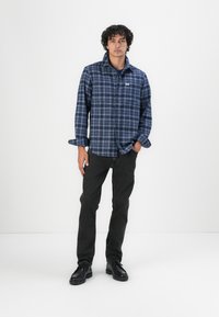 Camisa de franela a cuadros en tonos azul marino con mangas largas y botones al frente, combinada con jeans ajustados negros y zapatos negros de cordones.