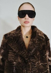 Cappotto in pelliccia sintetica marrone con un motivo testurizzato, abbinato a occhiali da sole neri di grandi dimensioni e una collana in oro con un ciondolo rotondo di pietra nera.