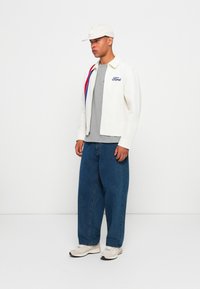 Young man wearing white cap, white Ford jacket, gray shirt, wide blue jeans, and white sneakers standing against plain white wall.