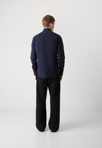 Cămașă albastră închis cu mânecă lungă, guler ascuțit și manșete cu nasturi, purtată cu pantaloni negri largi, văzută din spate pe un fundal simplu.