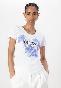 Bílá bavlněná tričko s krátkými rukávy, které má na přední straně modrou květinovou grafiku a trojúhelníkový design loga "GUESS".