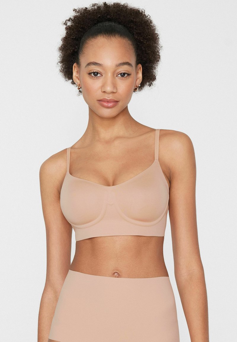 Mujer con cabello rizado, usando un sujetador beige sin costuras y ropa interior de talle alto a juego, mirando hacia adelante sobre un fondo blanco liso.