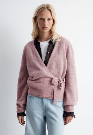 Light pink wrap cardigan made of soft material, featuring long sleeves, a tie at the waist, and a relaxed fit, layered over a dark button-up shirt.