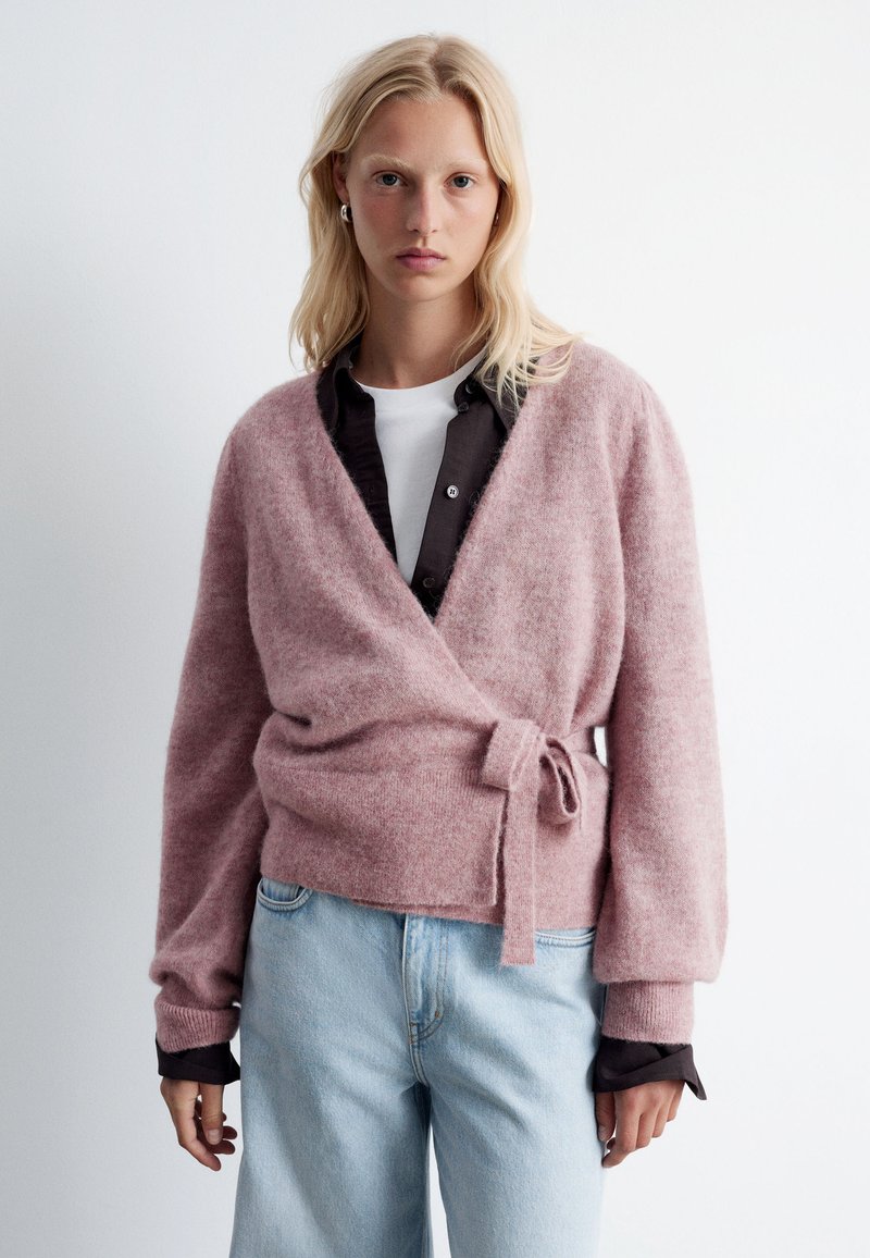 Light pink wrap cardigan made of soft material, featuring long sleeves, a tie at the waist, and a relaxed fit, layered over a dark button-up shirt.