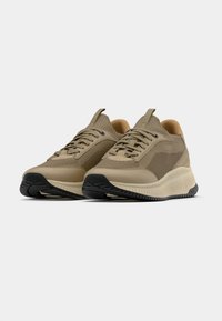 Ζευγάρι μπεζ αθλητικά παπούτσια από δίχτυ και δέρμα με χοντρές υφασμάτινες μπ εζ σόλες και μαύρο πέλμα, τοποθετημένα ελαφρώς προς τα αριστερά σε λευκό φόντο.