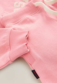 Sweatshirt rose plié avec des poignets côtelés, une taille côtelée, un cordon blanc et une étiquette noire portant la mention "Superdry" sur la couture latérale.
