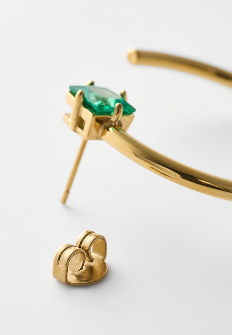 Pendiente de aro de oro con una piedra preciosa cuadrada verde y un cierre de mariposa de oro separado, descansando sobre una superficie blanca.