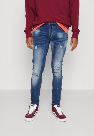 Osoba nosząca podarte niebieskie dżinsy, czerwone i białe trampki, koralową podkoszulkę i czerwony długi rękaw, stojąca na jednolitym tle.