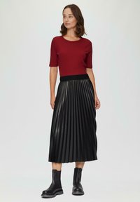 Rode geribbelde top met korte mouwen, gecombineerd met een zwarte geplooide rok met leren textuur en een elastische tailleband. Zwarte enkellaarsjes maken de outfit compleet.